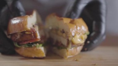 Siyah lastik eldivenli şefin elleri, burger ve sosun yarıya yakın çekimden akan yarımlara basıyor. Lezzetli yemek hazırlığı. Sulu iştah açıcı hamburger masada. Yavaş çekim.