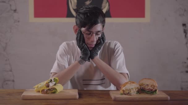Νέος αδύνατος άντρας με γυαλιά και μαύρα γάντια κάθονται στο τραπέζι μπροστά από ορεκτικό burger και το σαλάγμα επιλέγουν τι να φάνε πρώτα. Ο τύπος που δειπνά στο μοντέρνο εστιατόριο — Αρχείο Βίντεο