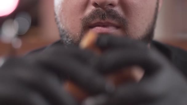 Närbild porträtt av en skäggiga mannen i svarta handskar äta en välsmakande hamburgare. Mannen njuter av läckra snabbmat i den moderna restaurangen. — Stockvideo