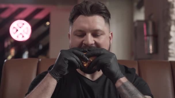 Retrato de un hombre barbudo con guantes negros comiendo una sabrosa hamburguesa sentada a la mesa en un sofá de cuero en la cafetería. El hombre que disfruta de la comida rápida en el restaurante moderno — Vídeo de stock