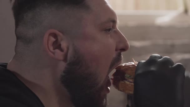 Боковой портрет бородатого человека в черных перчатках, поедающего вкусный бургер. Мужчина, наслаждающийся моющим рот фаст-фудом в современном ресторане . — стоковое видео