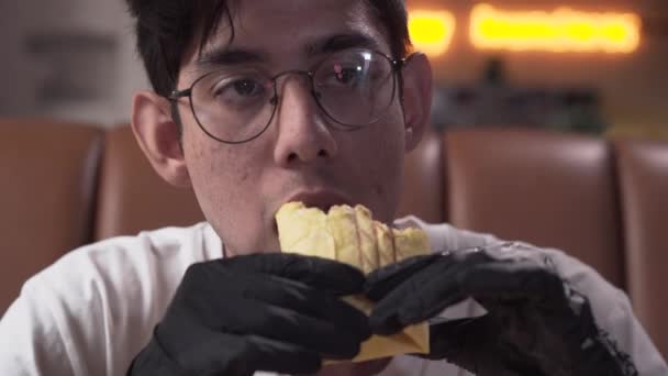 Portrét vyzáblého mladého muže v brýlích a černých rukavičkách, které jedí chutnou šawermu. Muž, který si užívá rychlého občerstvení v moderní restauraci — Stock video