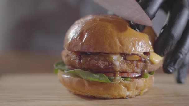 Handen van de chef-kok in zwarte rubberen handschoenen snijden hamburger met de grote scherpe mes close-up. Smakelijke voedselbereiding. Sappige overheerlijke Hamburger op tafel. Slow Motion. — Stockvideo