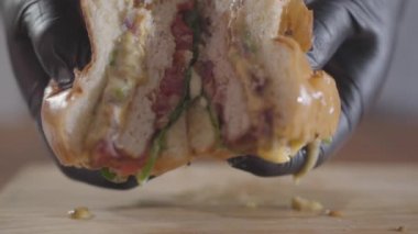 Siyah lastik eldivenli şefin yakın elleri burger ve sosun yarıya kadar bastırArak dışarı akıyor. Lezzetli yemek hazırlığı. Masada sulu ağız sulandıran burger. Yavaş çekim.
