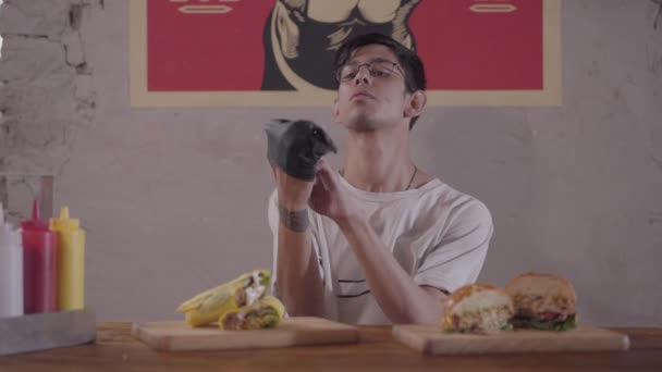 Hombre barbudo sentado en la mesa frente a una hamburguesa deliciosa y shawarma preparándose para comer. El tipo que se puso guantes negros . — Vídeos de Stock