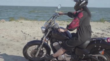 Siyah kask takan kız motosikletin üzerinde otururken deniz sahilinde uzağa bakıyor. Hobi, seyahat ve aktif yaşam tarzı. Bisikletçi dışarıda motosikletine biner. Motosikletle eğlence ve seyahat.