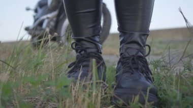 Siyah deri pantolon ve spor ayakkabı alanında çim üzerinde duran tanınmayan bir bisikletçinin bacakları. Motosikletin arka planında motosikletli. Hobi, seyahat ve aktif yaşam tarzı.