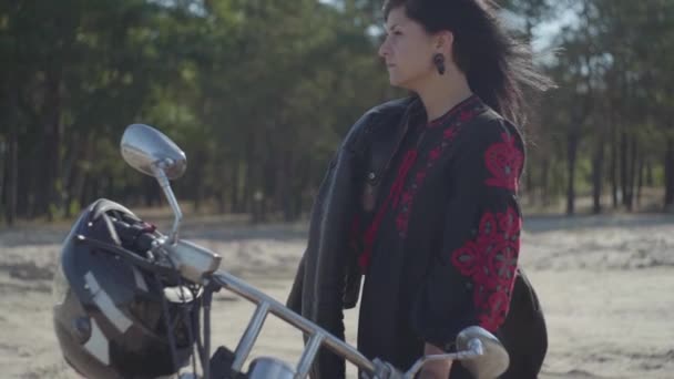 Jolie fille brune caucasienne debout à la moto regardant loin devant la forêt de pins. Hobby, voyage et mode de vie actif. Attrayant motard féminin sur sa moto à l'extérieur. Mouvement lent . — Video