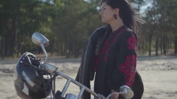 Linda chica morena caucásica de pie en la motocicleta mirando hacia otro lado frente al bosque de pinos. Hobby, viajar y estilo de vida activo. Atractiva motorista femenina en su moto al aire libre. Movimiento lento . — Vídeo de stock