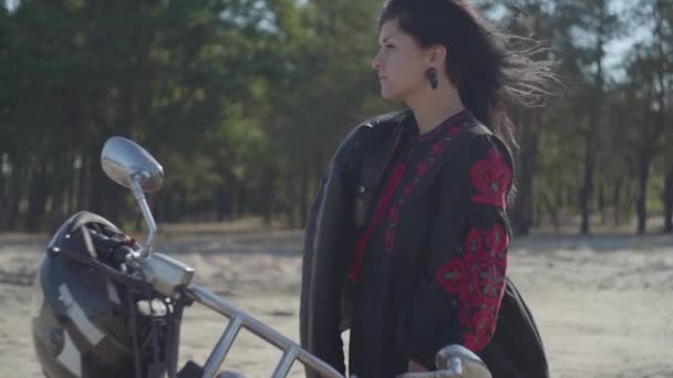 Dziewczyna z czarnym włosami stoi na motocyklu patrząc z dala przed lasem sosnowym. Hobby, Podróżowanie i aktywny tryb życia. Atrakcyjna kobieta motocyklista na zewnątrz na motocyklu. Zwolnionym. — Wideo stockowe