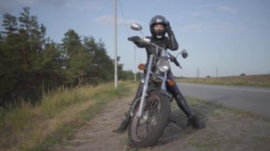 Yolda bisikletinin üzerinde oturan deri elbiseli başarılı kendine güvenen genç kadının ön görünümü. Hobi, seyahat ve aktif yaşam tarzı. Kadın motorcu motosikletinin üzerinde dışarıda. Yavaş çekim.