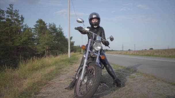 Vista frontal de la joven confiada en vestido de cuero sentada en su bicicleta en la carretera. Hobby, viajar y estilo de vida activo. Motociclista al aire libre en su moto. Movimiento lento . — Vídeo de stock