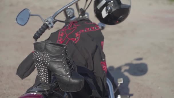 Spiked buty z wysokimi obcasami i czarno-czerwona sukienka leżąca na motocyklu zbliżenie. Motocykl z hełmem na kole na brzegu rzeki. Hobby, Podróżowanie i aktywny tryb życia — Wideo stockowe