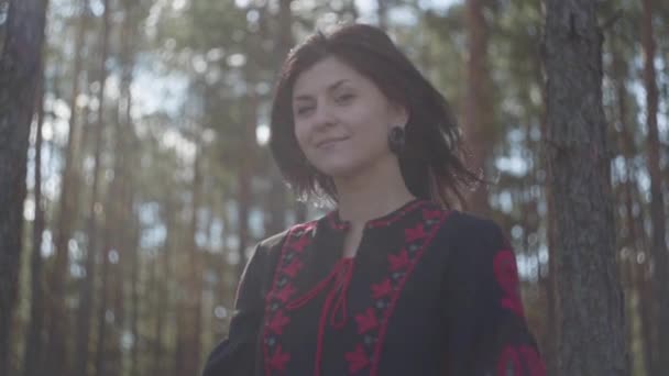 Linda mujer joven caucásica en vestido negro y rojo de pie en el bosque de pinos mirando a la cámara. Conexión con la naturaleza. Movimiento lento . — Vídeos de Stock