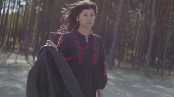 Vonzó kaukázusi fiatal nő, gyönyörű, hosszú, fekete és piros ruhában állva a fenyőerdőben. Kapcsolat a természettel. Lassított mozgás. — Stock videók