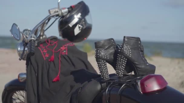 Zapatos de punto con tacones altos y vestido negro y rojo acostado en el primer plano de la motocicleta. La moto con el casco en la rueda en la orilla del río. Aficion, viajes y estilo de vida activo — Vídeos de Stock