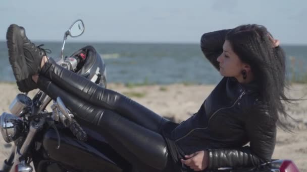 Mujer caucásica bastante joven en una chaqueta de cuero negro y pantalones acostados en una motocicleta cerca del mar o el río. Hobby, viajar y estilo de vida activo. Ocio y viajes en moto . — Vídeo de stock