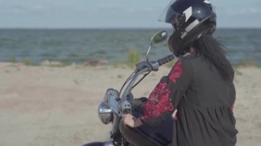 Siyah elbise ve kask giyen beyaz kız motosikletin üzerinde otururken kameraya bakıyor. Hobi, seyahat ve aktif yaşam tarzı. Bisikletçi dışarıda motosikletine biner. Motosikletle eğlence ve seyahat.