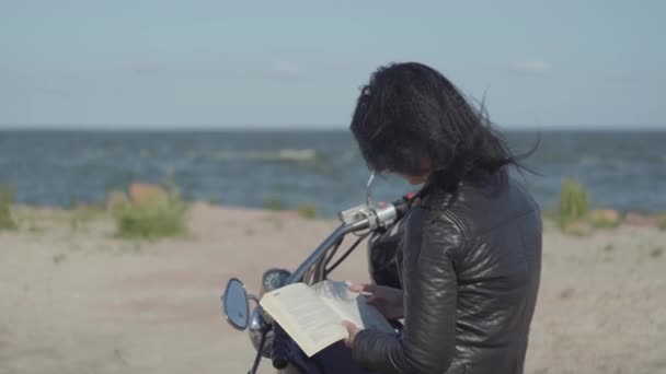 Vista posteriore della bella ragazza caucasica con una giacca di pelle nera seduta sul libro di lettura della moto. Hobby, viaggiare e stile di vita attivo. Tempo libero e viaggi in moto . — Video Stock