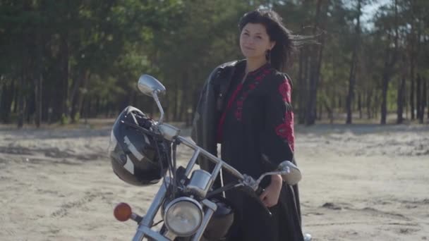 La chica con el pelo negro de pie en la motocicleta mirando a la cámara delante del bosque de pinos. Hobby, viajar y estilo de vida activo. Atractiva motociclista al aire libre en su moto. Despacio. — Vídeos de Stock