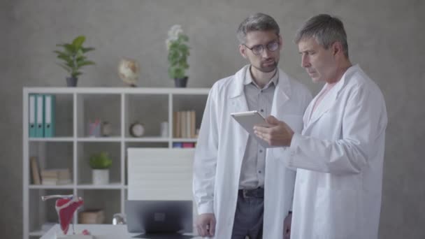 Dva mužští lékaři kontrolují informace na tabletu a diskutují o nich. Koncept medicíny, techniky, zdravotnictví a lidí, nemocnice. Nové moderní plně funkční lékařské zařízení. — Stock video