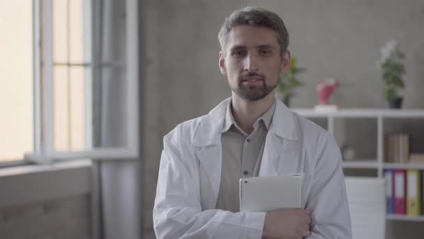 Porträtt av stilig skicklighet leende man i vit dräkt ser i kameran håller tabletten. Vuxen läkare i modern privat klinik. Begreppet medicinsk behandling — Stockvideo