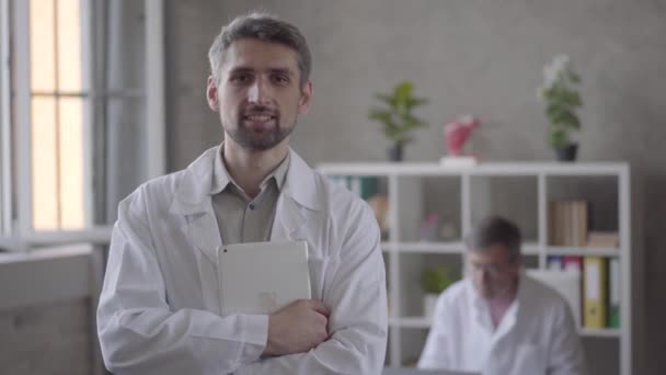 Porträt eines gut aussehenden lächelnden Arztes, der in die Kamera blickt und Tablet auf den Hintergrund seines Kollegen in der Klinik hält. neue moderne, voll funktionsfähige medizinische Einrichtung. — Stockvideo