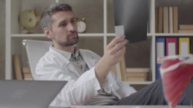 Usta erkek doktorlar modern kliniğin ofisinde röntgen ikontrol. Beyaz cüppeli genç adam çalışma masasında bir sandalyede oturan x-ray görünümlü. Yeni modern tam fonksiyonel tıbbi tesis.