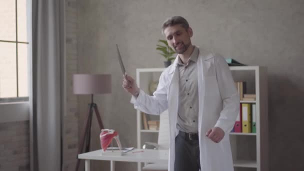 Bel homme positif, réussi et satisfait en robe blanche dansant avec rayons X dans les mains dans la clinique moderne. Le docteur a eu de bonnes nouvelles, il est heureux. Concept de médecine, soins de santé, hôpital . — Video