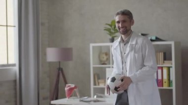 Yakışıklı olumlu, başarılı ve memnun adam beyaz cüppe modern klinik ofisinde topla oynuyor. Mutlu doktor harika haberler aldı. Tıp kavramı, sağlık, hastane.