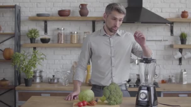 Guapo hombre bien parecido en la camisa de pie en la mesa con una licuadora que se prepara para hacer un batido de verduras. Concepto de comida saludable — Vídeo de stock