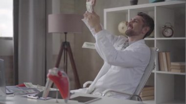 Yakışıklı mutlu başarılı, memnun sakallı doktor klinik ofisinde bir çalışma masasında bir sandalyede oturan dolar dağılım. Para yağmuru, düşen dolarlar. Yavaş çekim.