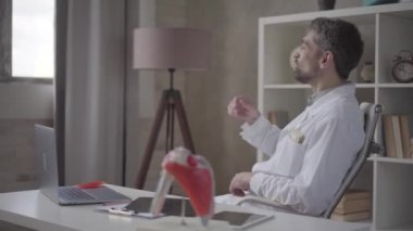 Mutlu başarılı, memnun sakallı doktor klinik ofisinde bir çalışma masasında bir sandalyede oturan dolar dağılım. Para yağmuru, düşen dolarlar. Yavaş çekim.