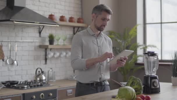 Szép csinos férfi az ing főzés vacsora ellenőrzése recept a tablettát a konyhában otthon. Fogalma az egészséges táplálkozás, az otthoni főzés. A fickó főzés élelmiszer munka után — Stock videók