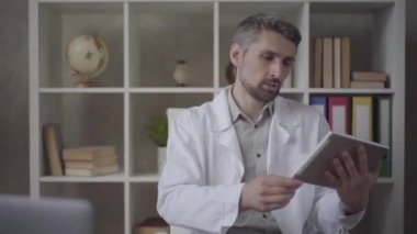 Beyaz cüppe portre çekici adam akciğerx-ray resmi ile tablet gösteren kamera bakıyor. Modern özel klinikte usta doktor. Yeni tam işlevsel bir sağlık tesisi. Yavaş çekim.