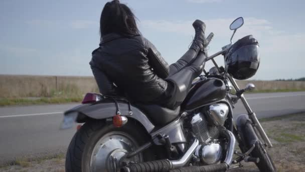 黒い革のジャケットと古典的なオートバイに横たわるズボンのかわいい白人女性。趣味、旅行、アクティブなライフスタイル。バイクでのレジャーと旅行. — ストック動画