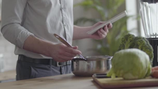Невпізнаваний чоловік у сорочці рецепт перевірки супу на планшеті на кухні вдома. Концепція здорової їжі, домашньої кухні. Хлопець готує їжу після роботи — стокове відео