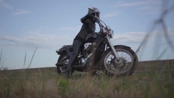 Jolie femme caucasienne dans une veste en cuir noir et casque chevauchant une moto classique. Fille enlève le casque debout près de son vélo sur le bord de la route. hobby, voyage et mode de vie actif . — Video