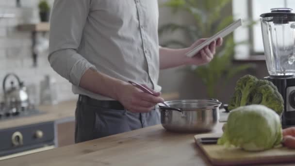 Ο αγνώριστο όμορφος άντρας με το μπλουζάκι που μαγειρεύει σούπα, ελέγχει τη συνταγή του δισκίου στην κουζίνα στο σπίτι. Η ιδέα της υγιεινής διατροφής, το σπιτικό μαγείρεμα. Ο τύπος που μαγειρεύει φαγητό μετά τη δουλειά. — Αρχείο Βίντεο