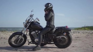 Siyah deri ceket ve kask motosiklet üzerinde oturan sevimli beyaz kadın. Kız kask kaldırır ve saç düzleştirmek. Hobi, seyahat ve aktif yaşam tarzı. Motosikletle eğlence ve seyahat.