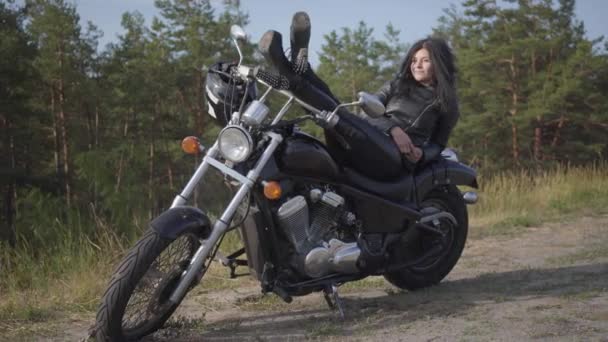 Bedårande ung kaukasiska kvinna i en svart skinnjacka och byxor som ligger på en klassisk motorcykel. Hobby, resande och aktiv livsstil. Fritid och resa med motorcykel. — Stockvideo