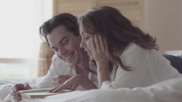 Boldog fiatal pár feküdt az ágyon olvasatban a könyv közelről. Szép nő és férfi kiadási időt az ágyban otthon. Fogalma boldogság és a gyengédség, az emberek a szeretetben. — Stock videók