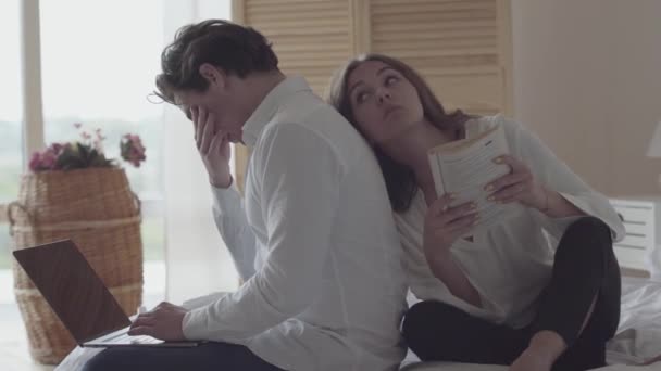 Joven cansado trabajando en el portátil en casa o en el hotel mientras su esposa leía el libro junto a él. Familia joven pasando tiempo juntos. Diferentes estilos de vida y aficiones. Ocio interior — Vídeos de Stock