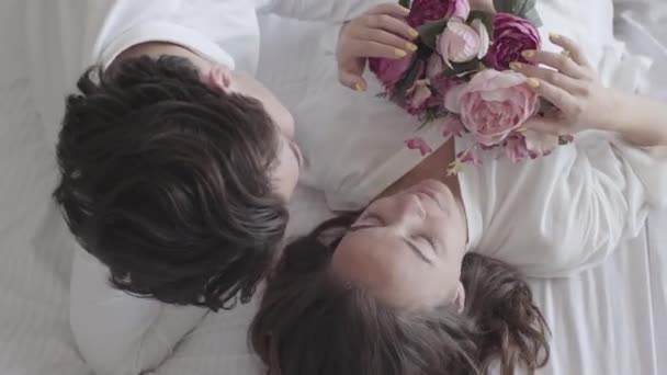 Overhead shot happy young couple lying on the bed talking, woman holding bouquet smiling close-up. Tender relationship concept, people in love. Top view — Stock Video