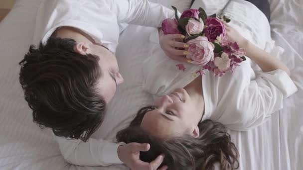 Overhead shot happy young couple lying on the bed talking, woman holding bouquet smiling close-up. Tender relationship concept, people in love. Top view. Slow motion. — Stock Video