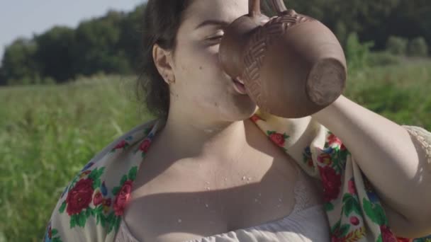 Mooie Mollige vrouw geniet van het drinken van verse melk van de aarden kruik op het groene zomer veld. Prachtig landschap. Folklore, tradities concept. Echte landelijke vrouw. Slow Motion — Stockvideo