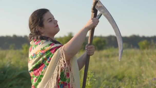 Yeşil yaz alanında biçme başlatmak için hazırlanıyor tırpan keskinleştirme güzel kilolu kadın portresi. Çiftçi tırher'ı öğütücüyle bileyor. Folklor, gelenekler kavramı. Sahada çalışma — Stok video