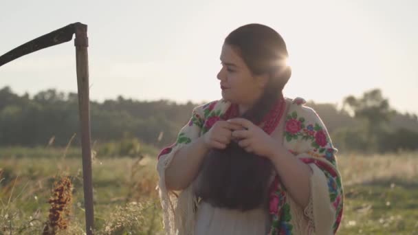 Hermosa mujer con sobrepeso con una guadaña trenzándose el pelo a la luz del sol en el verde campo de verano. Folclore, concepto de tradiciones. Trabaja en el campo. Lente bengalas. Mujer rural real . — Vídeos de Stock