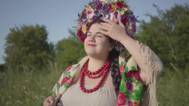 Retrato de una linda mujer regordeta con una corona en la cabeza sonriendo a la luz del sol en el verde campo de verano. Conexión con la naturaleza. Mujer rural real . — Vídeo de stock