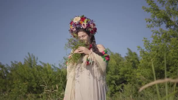 Schöne übergewichtige Frau mit einem Kranz auf dem Kopf, die auf der grünen Sommerwiese Blumen schnüffelt und dann einem Mann in Arbeitskleidung hinterherläuft. Landleben. Flirt-Konzept. Echte Landfrau. — Stockvideo